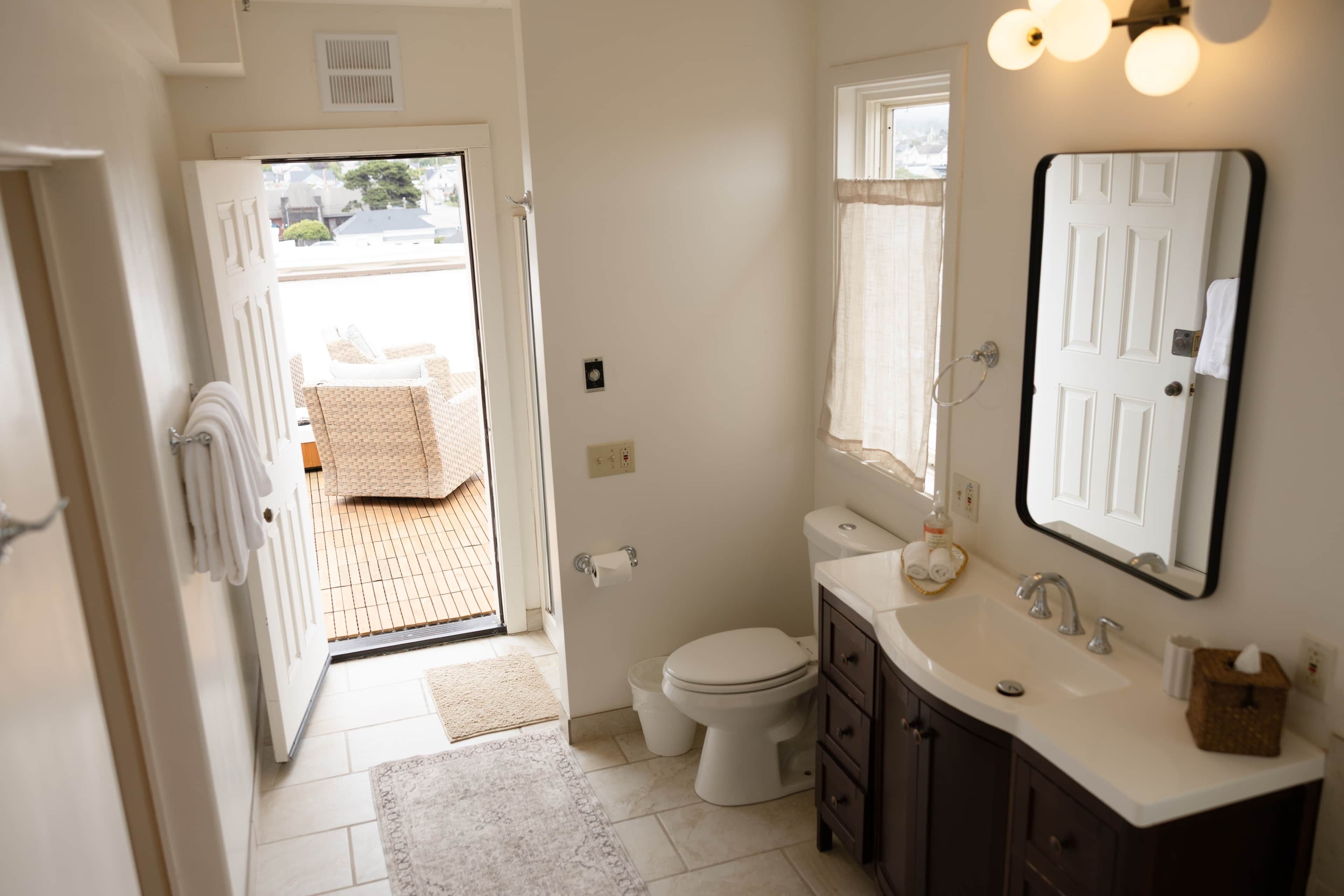 Une salle de bain équipée de toilettes, d’un lavabo et d’un miroir. Une porte donne sur un espace extérieur où se trouvent une chaise et une serviette.