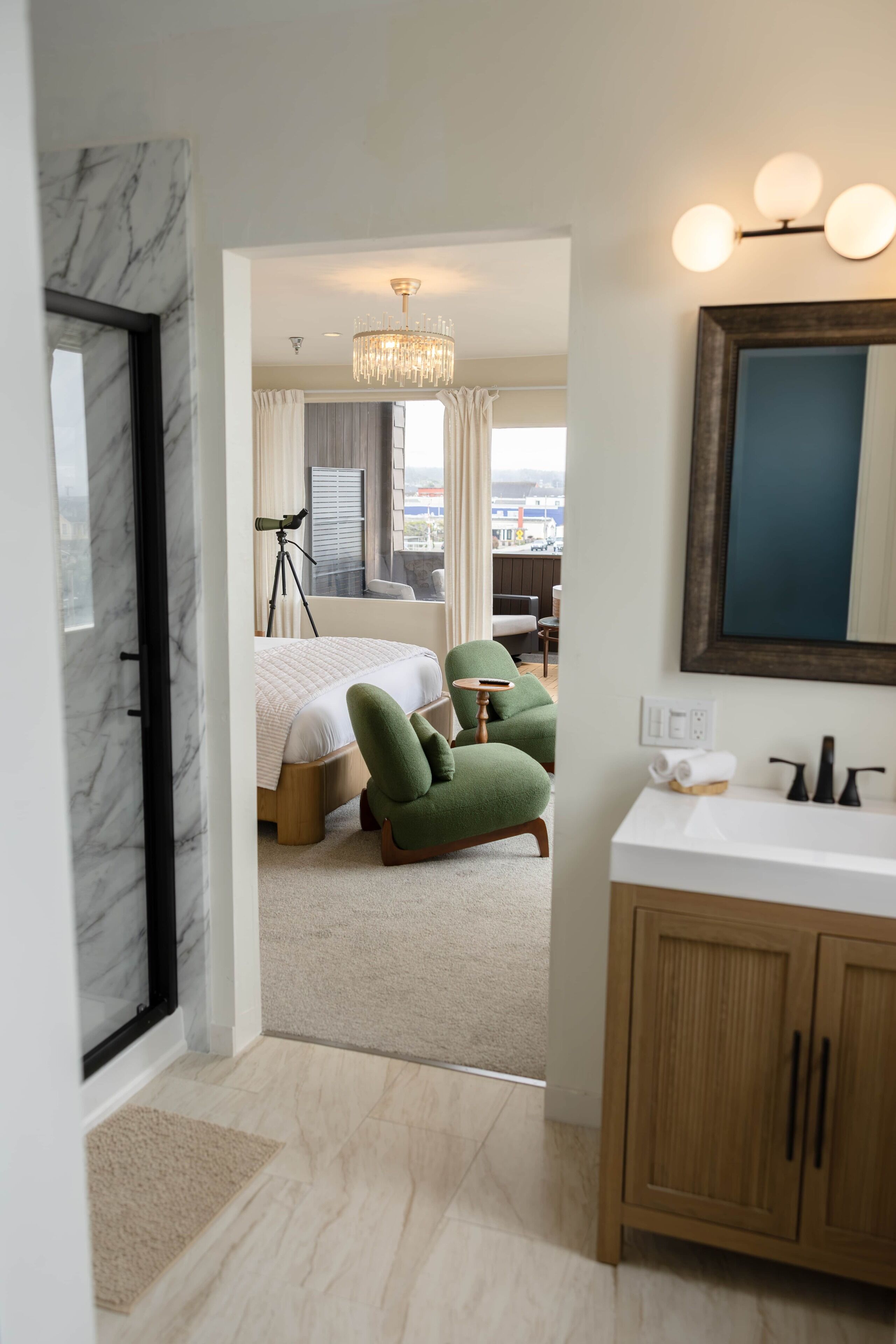 Ein Hotelzimmer mit einem Bett, einem grünen Sessel, einem Schreibtisch, einer Chandelierleuchtre und einem Badezimmer mit Spiegel und Waschbecken.