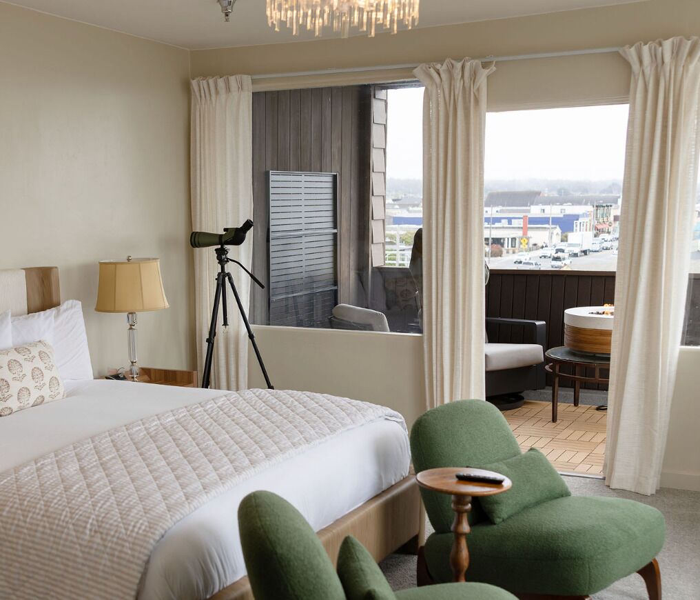 Een hotelkamer met een bed, een bureau, een stoel, een lamp, een telescoop en uitzicht op de stad.