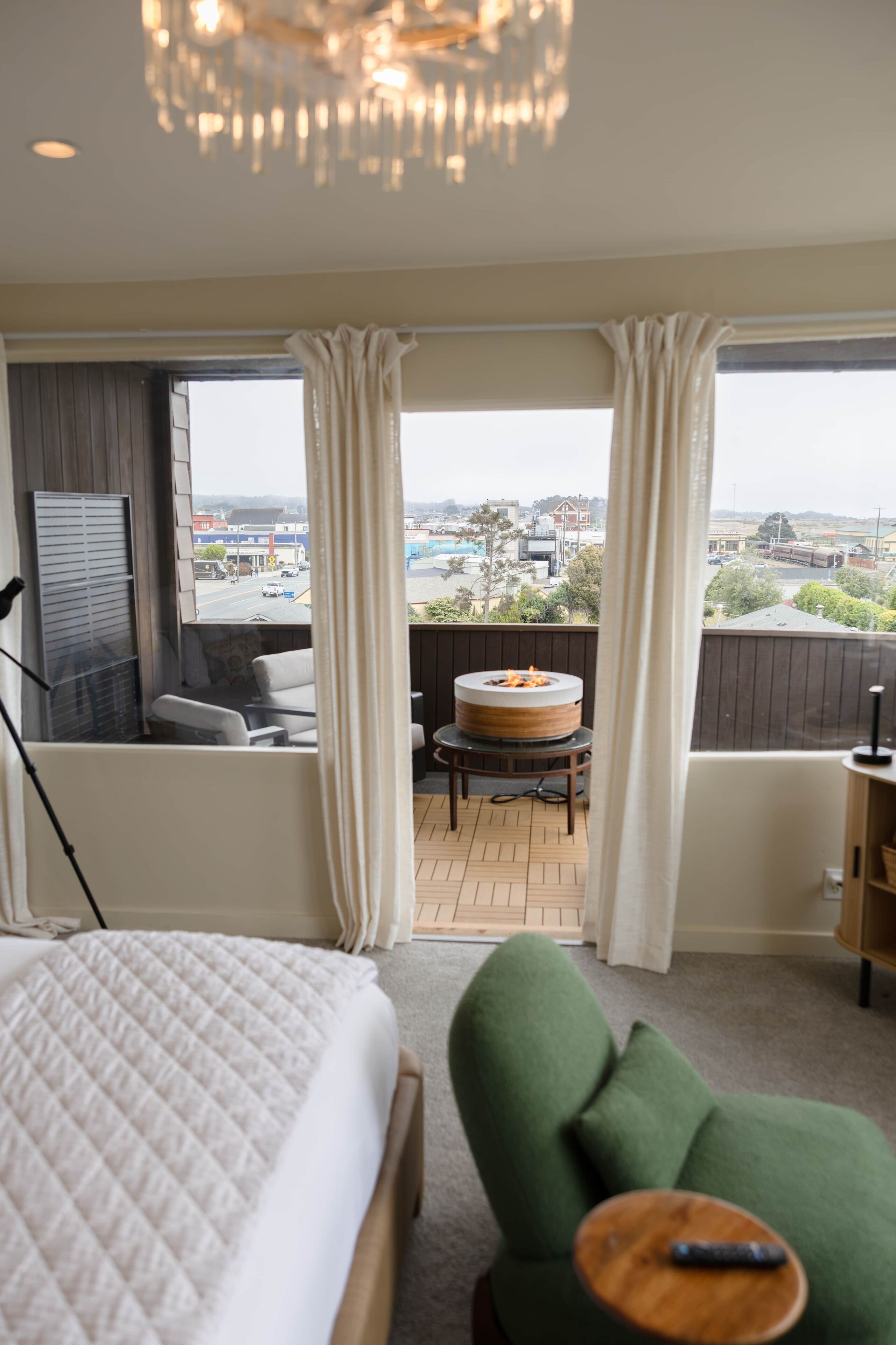 Un dormitorio con cama, un sillón verde, una pequeña mesa redonda, una chimenea y vistas de la ciudad a través de amplios ventanales.