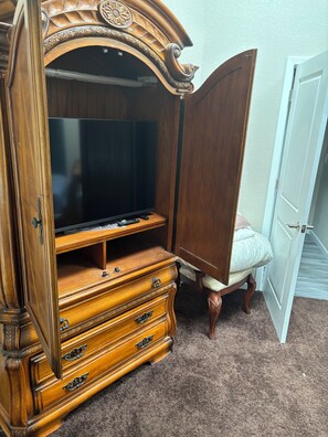 Ein Holzschrank mit Fernseher und Fernbedienung, ein Sessel mit Kissen und ein Teppichboden.