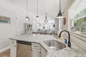 A kitchen with a granite countertop, stainless steel sink, and a dishwasher. There is a dining area in the background with a chandelier. - 1 Block to Beach | New Outdoor Living Space (St Augustine)