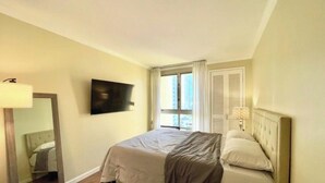 A hotel room with a bed, a flat-screen TV mounted on the wall, a window with curtains, and a bedside lamp. - Perfect Ocean view 2-bedroom condo that’s centrally located near Ala Moana beach (Honolulu)
