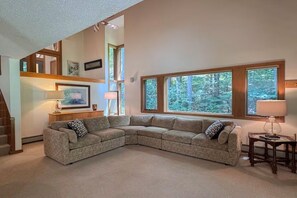 A living room with a sectional sofa, a small table, and a lamp. There are large windows with a view of trees. - Mountainside Retreat @ Attitash, Ski-In, Ski-Out, Pool, Hiking (Bartlett)