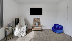 A room with a flat-screen TV mounted on the wall, a blue bean bag chair, a white chair with a textured blanket, and a small table with a statue and a mosaic piece. - Spacious Vegas Retreat • Close to Strip • Sleep 12 (Las Vegas)