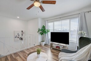 Un salon moderne avec un ventilateur de plafond, un téléviseur à écran plat, une plante en pot, une table basse ronde et un fauteuil gris.