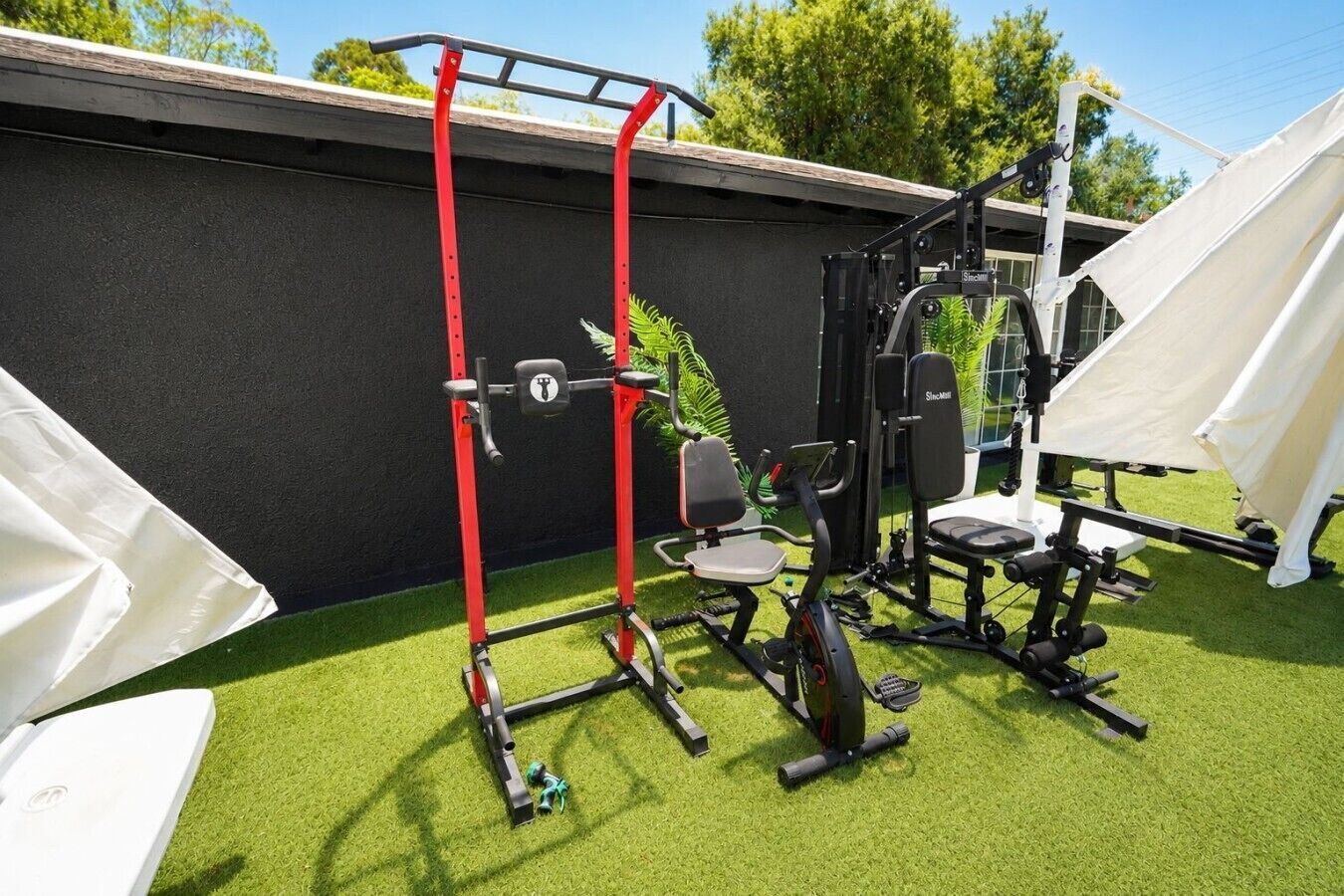 Ein Outdoor-Fitnessbereich mit Trainingsgeräten, darunter eine Klimmzugstange, eine Gewichtbank und ein Ergometer, vor dem Hintergrund eines Gebäudes und Bäumen.