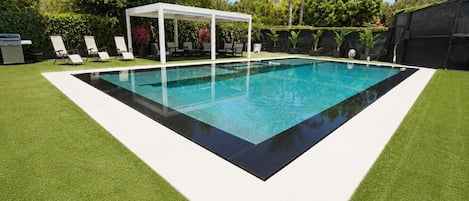 A rectangular swimming pool with a white deck and a covered seating area.