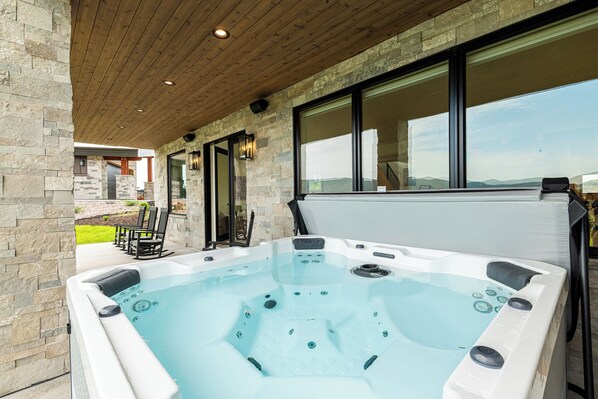 A hot tub with a wooden ceiling and stone walls, a patio with chairs, and a view of the landscape.