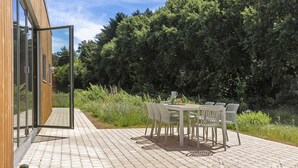 En udendørs terrasse med et bord og stole, en træbygning med glasdøre og en frodig grøn hæk i baggrunden.