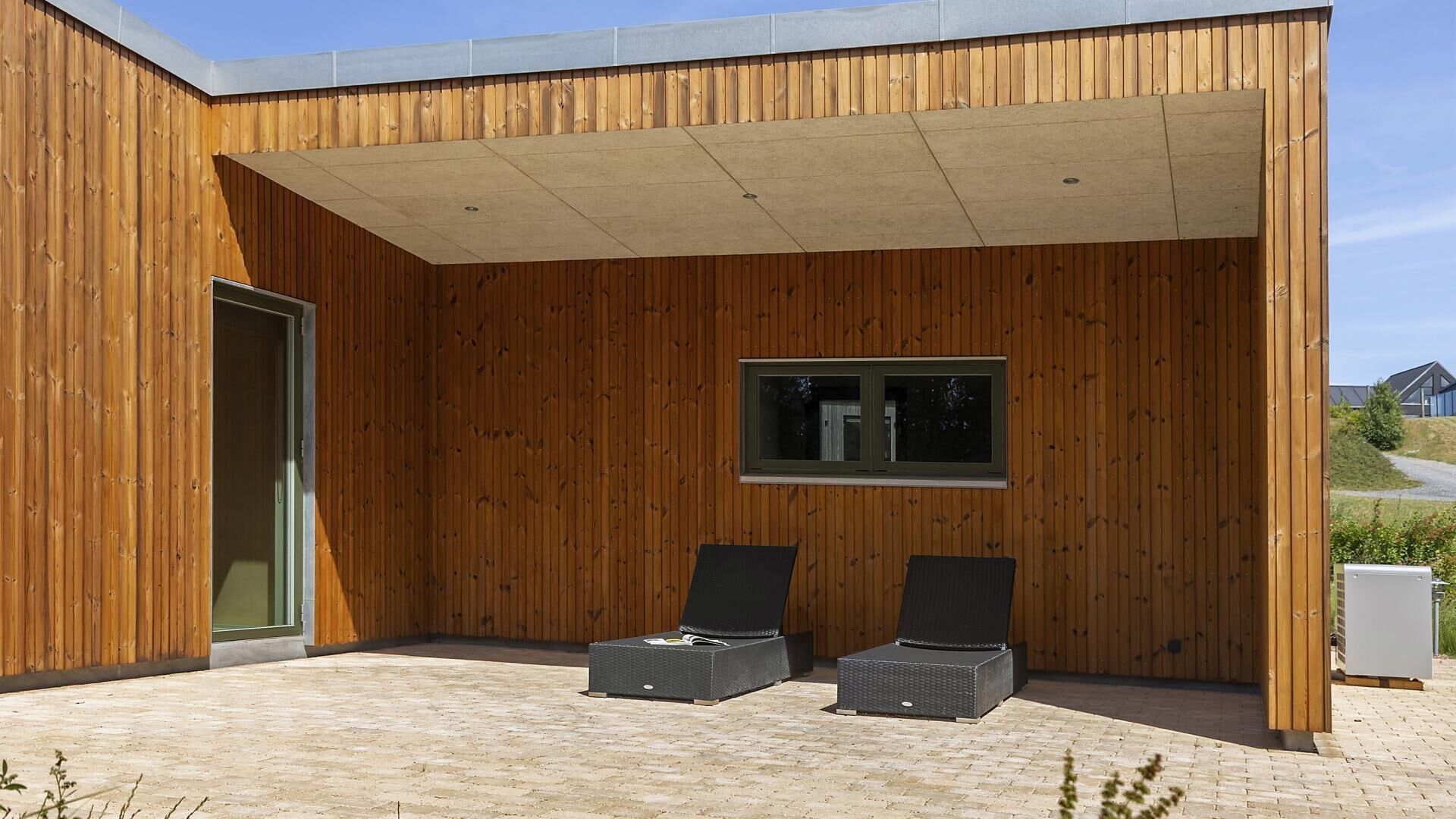 Ein modernes Gebäude mit Holzfassade, einer überdachten Terrasse mit zwei Liegestühlen und einem Fenster.