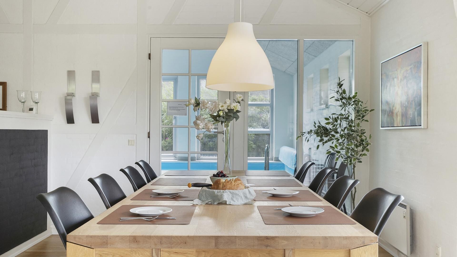 Een eetkamer met een grote tafel, stoelen, een hanglamp, een vaas met bloemen en uitzicht op de buitenlucht door een glazen deur.