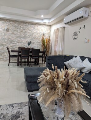 A living room with a stone wall, a dining area with a table and chairs, and a black sofa with a decorative plant. - Delightful 3-bedroom house with AC in charming Cunupia (Cunupia)