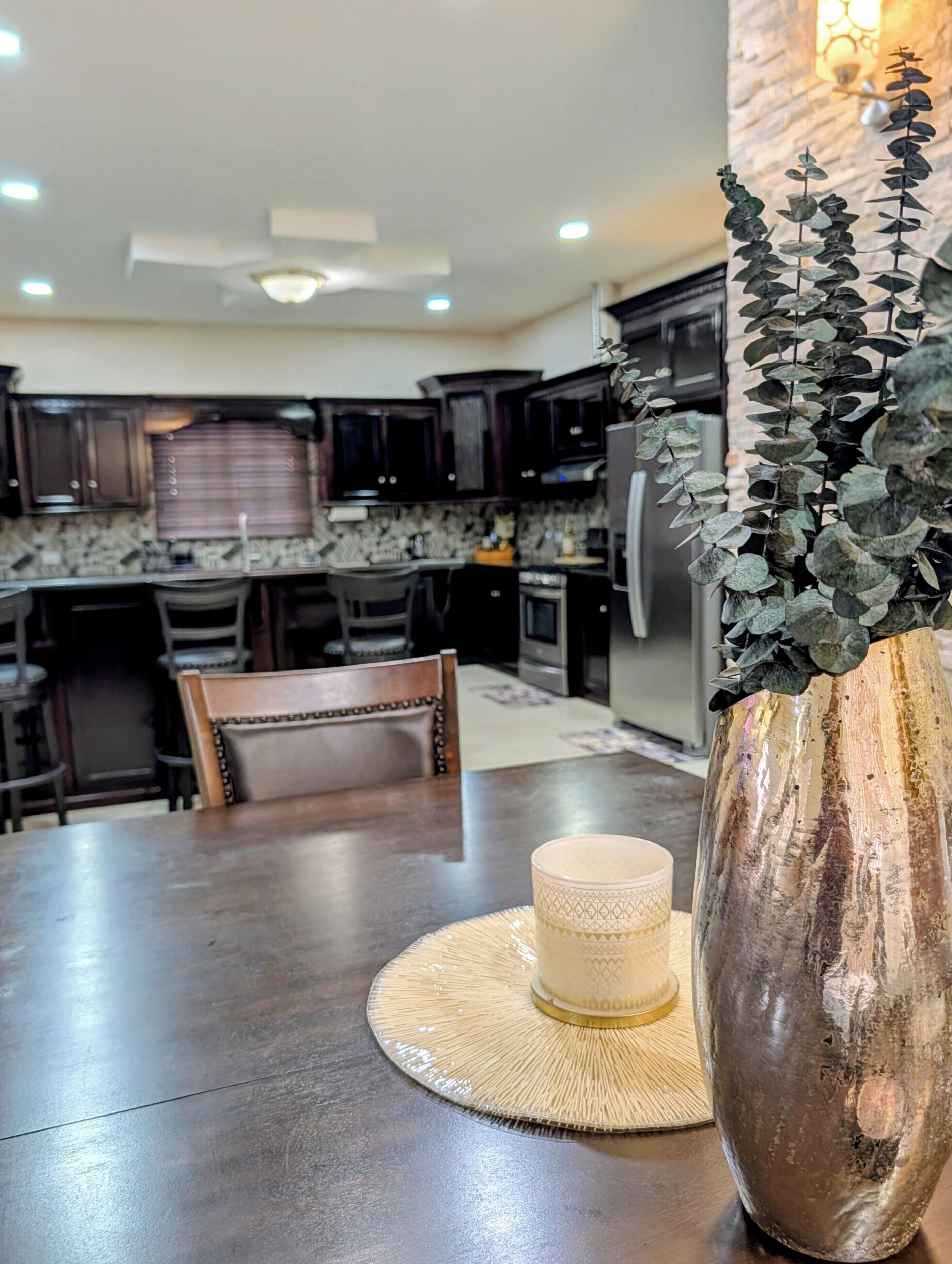 A kitchen with dark wooden cabinets, a dining table with a woven placemat, and a decorative vase with foliage.