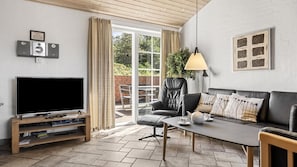 A modern living room with a flat-screen TV, a leather sofa, a dining table, and a sliding glass door leading to an outdoor patio. - VillaVilla 989 - Søndervig, Vestjylland (Ringkøbing)