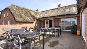 Eine Terrassenanlage mit Tischen und Stühlen, einem Reetdach und einem Backsteinhaus mit großen Fenstern.