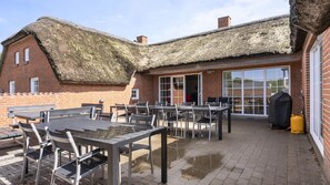 Eine Terrassenanlage mit Tischen und Stühlen, einem Reetdach und einem Backsteinhaus mit großen Fenstern.