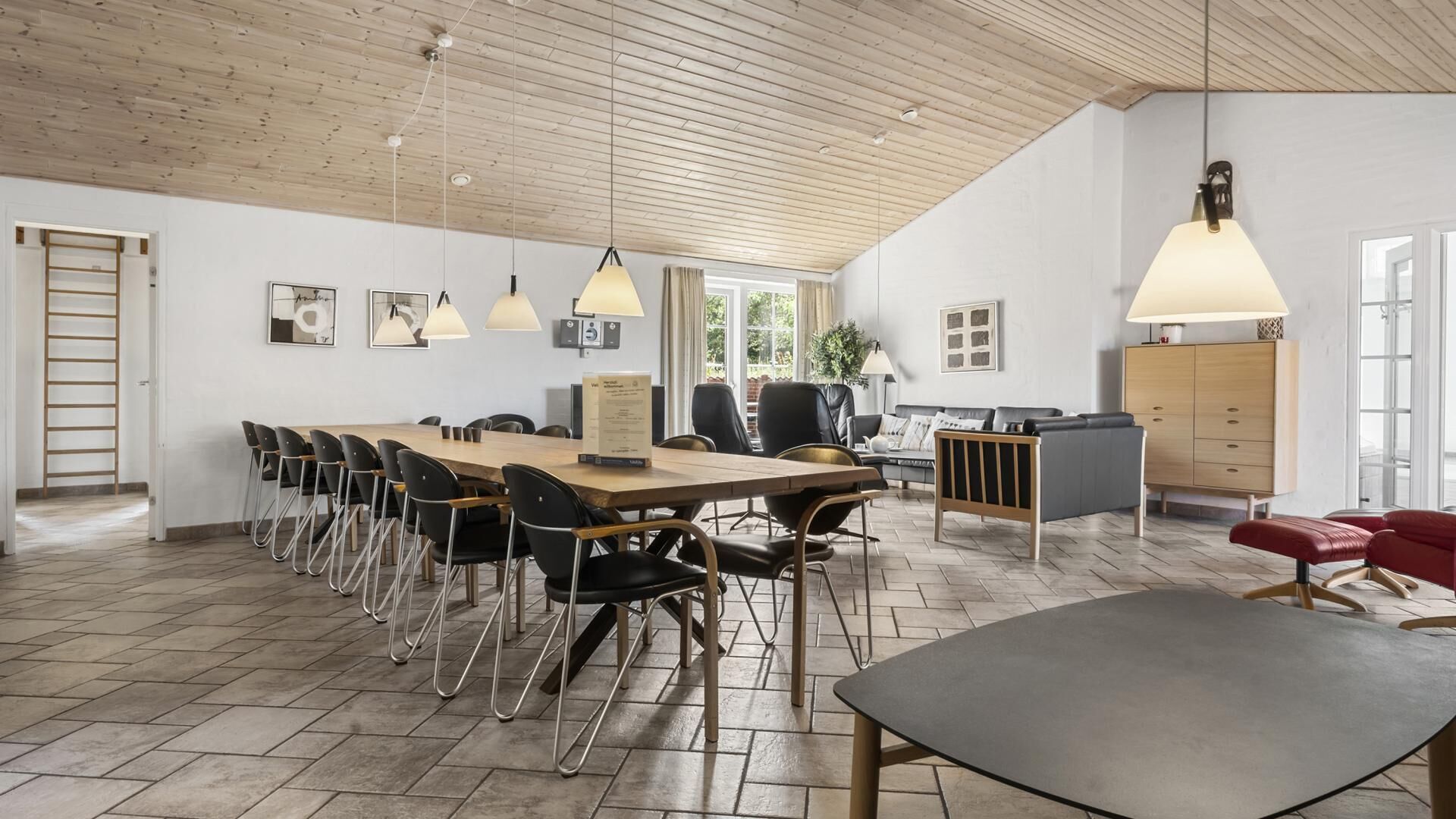 A spacious room with a long wooden table, black chairs, a grey sofa, and a red bench. There are pendant lights, a cabinet, and a ladder against the wall.