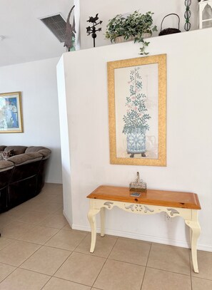 Una habitación con pared blanca, una obra de arte enmarcada de un jarrón con flores, una consola de madera, un sofá con cojines y un respiradero en el techo.