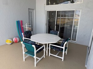 Uma área de varanda coberta com uma mesa e cadeiras brancas, uma porta de vidro deslizante e um pequeno espaço de recreação com suporte para bolas e uma rede.