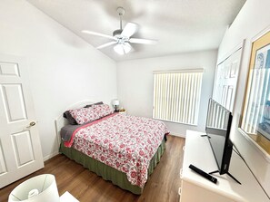 Ein Schlafzimmer mit einem Bett, einem Deckenventilator, einem Fenster mit Jalousien, einem Schreibtisch mit Computer und einer Tür.