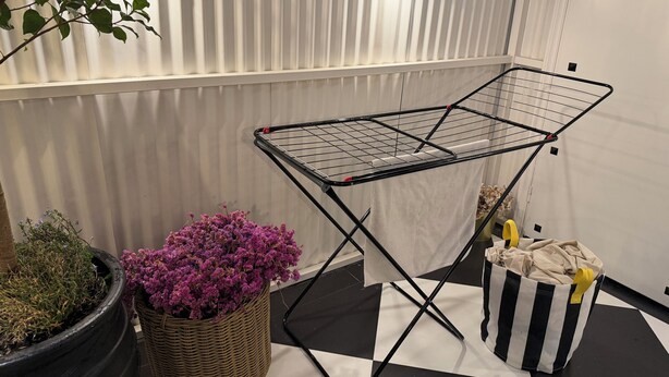 Een kamer met een zwart-wit geruite vloer, een zwarte metalen droogrek, een bloempot met paarse bloemen en een mand met een beige handdoek.