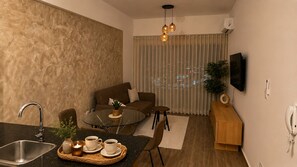 A modern living room with a sofa, a glass coffee table, a small dining table with cups, a kitchen area with a sink, and a TV mounted on the wall. - Modern Ocean View Oasis, Pool & Free Parking (Santo Domingo Este)
