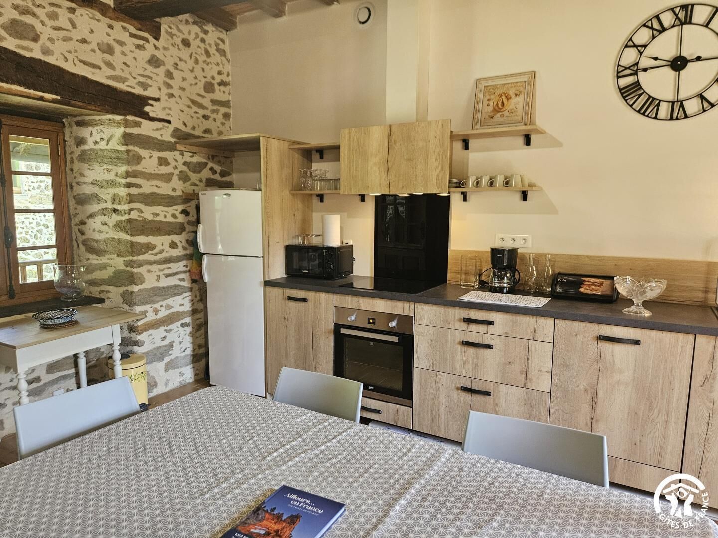 Une cuisine chaleureuse avec un mur en pierre, des armoires en bois, un réfrigérateur noir, un four à micro-ondes, une machine à café et une horloge au mur.