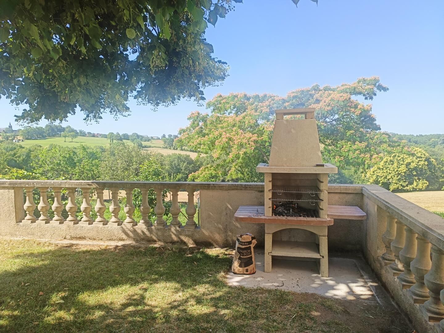 Een buitenbarbecue met een stenen schoorsteen, omgeven door een stenen balustrade, met op de achtergrond glooiende heuvels en bomen.