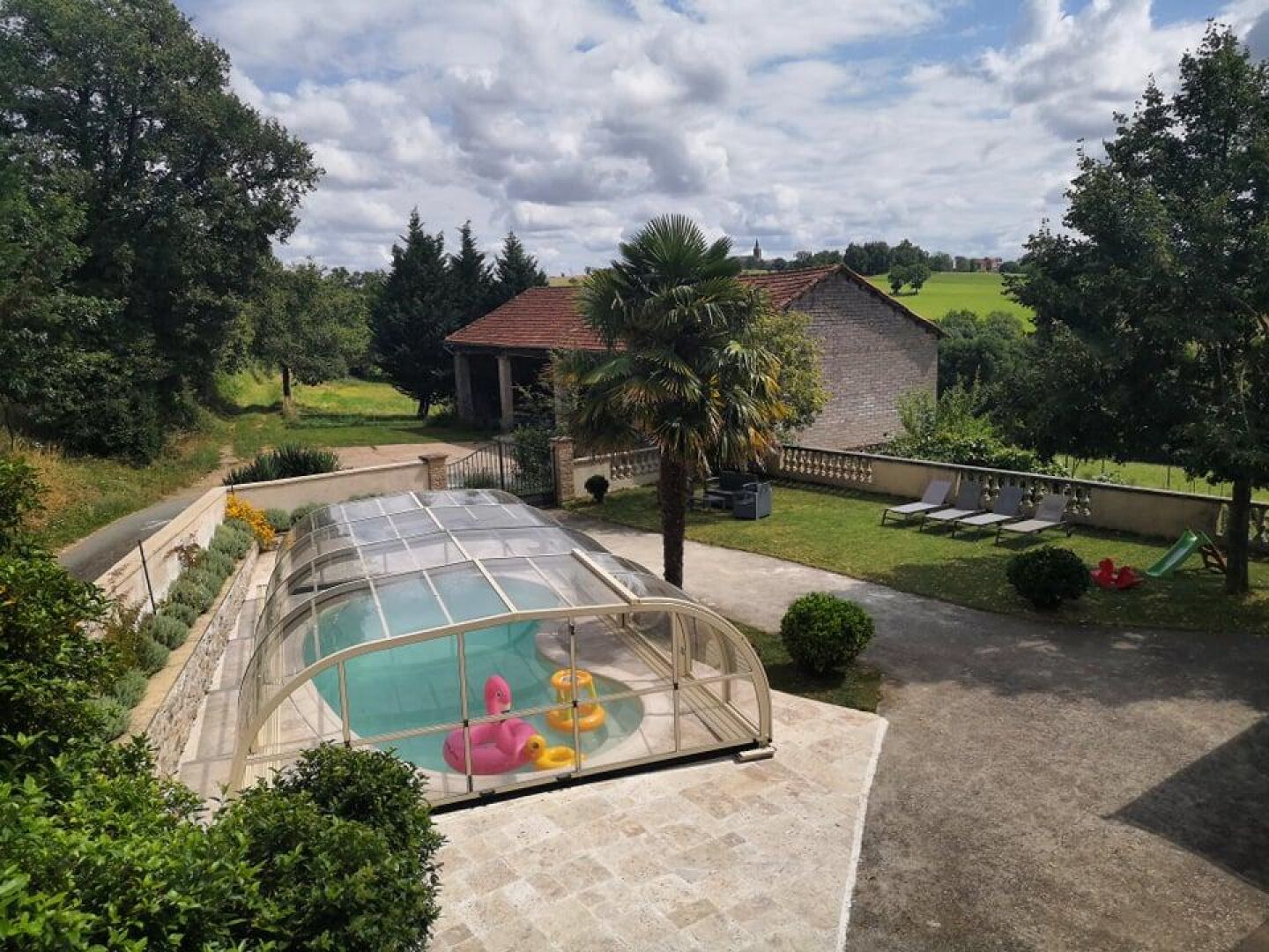 A swimming pool with a glass enclosure, surrounded by a landscaped garden with a brick building in the background.