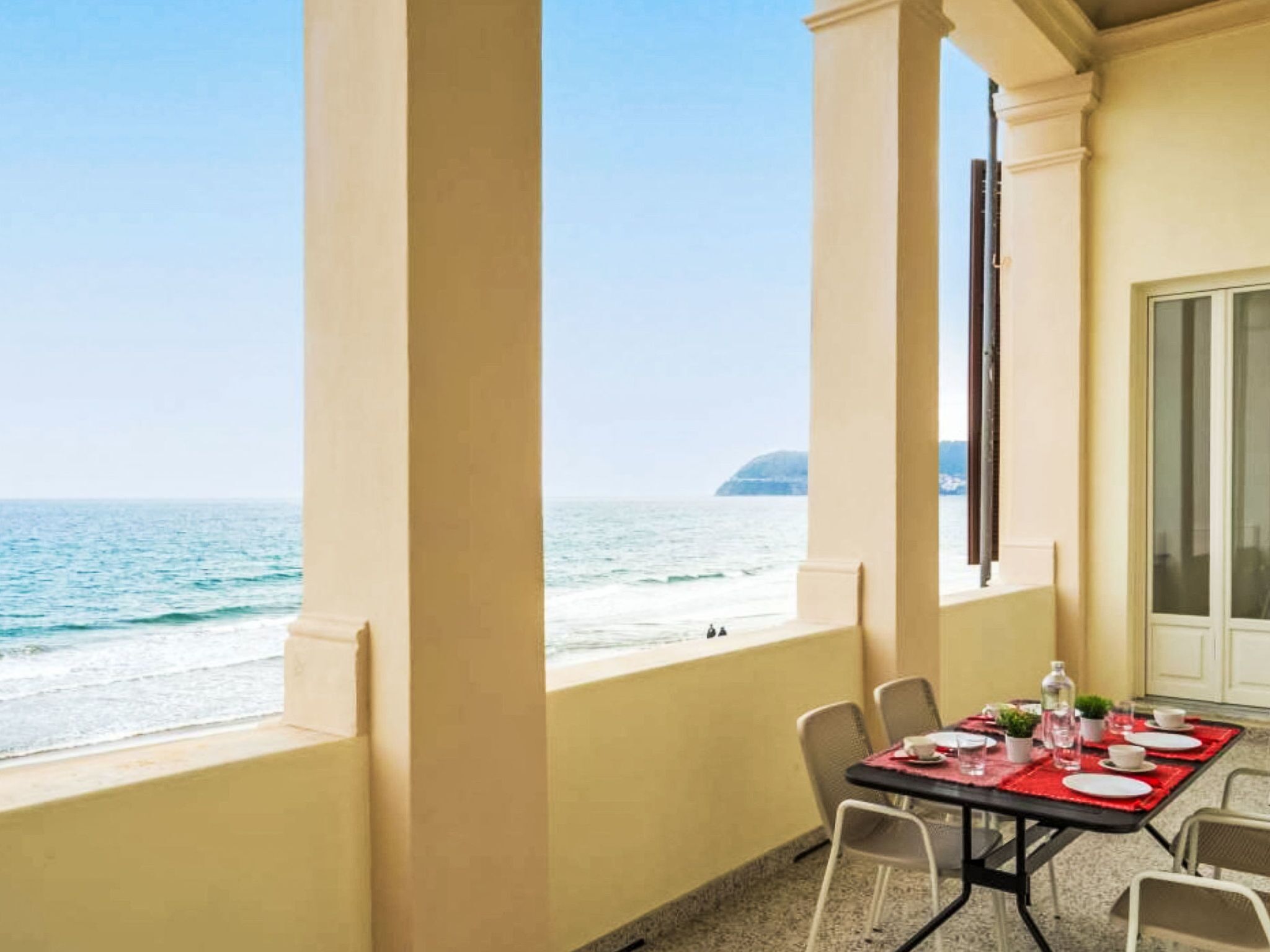 A balcony with a table set for dining, overlooking a beach with the ocean and a distant island.