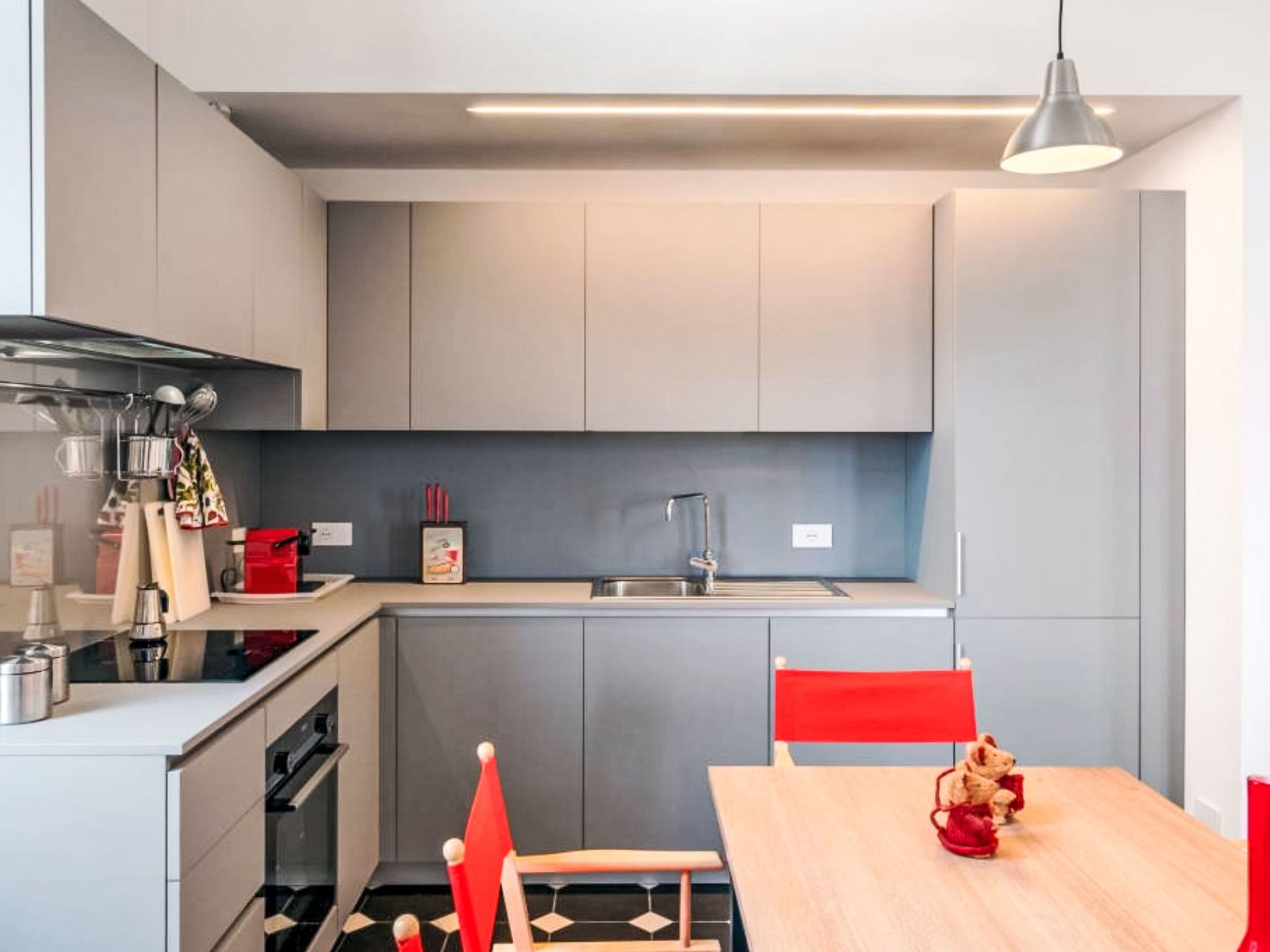 A modern kitchen with grey cabinets, a stainless steel sink, and a wooden dining table with red chairs.