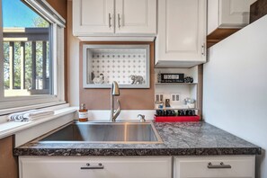 A kitchen with a stainless steel sink, white cabinets, a marble countertop, and a window with blinds. - Cozy Garden Retreat | Pet Friendly w/ Private Yard (Colorado Springs)
