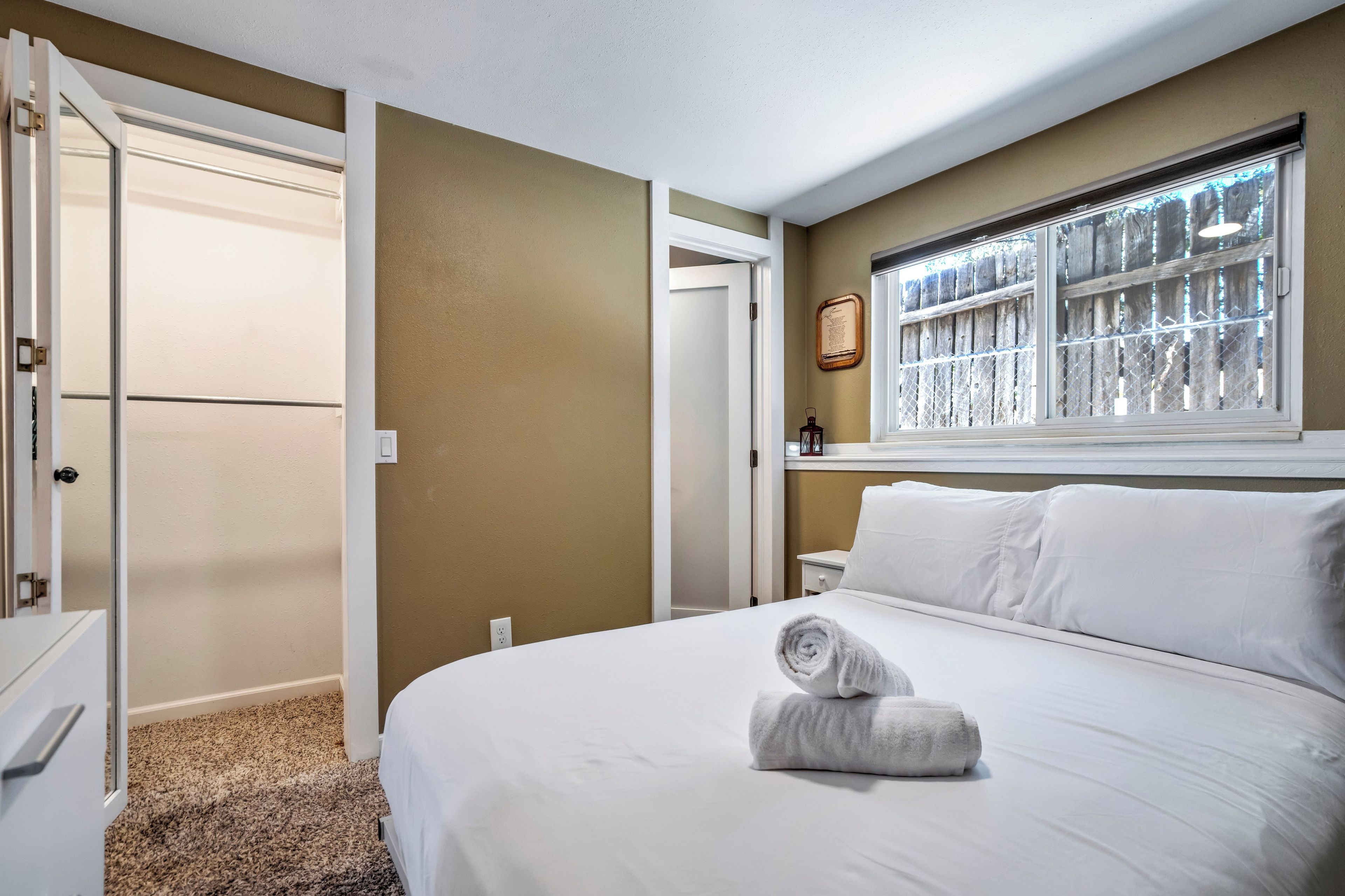 Habitación de hotel con una cama, ventana con vista a una valla de madera, una puerta y un aplique en la pared.