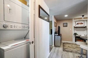 A laundry room with a front-loading washer and dryer, a hallway leading to a living area with a fireplace, and a mounted mirror. - Cozy Garden Retreat | Pet Friendly w/ Private Yard (Colorado Springs)