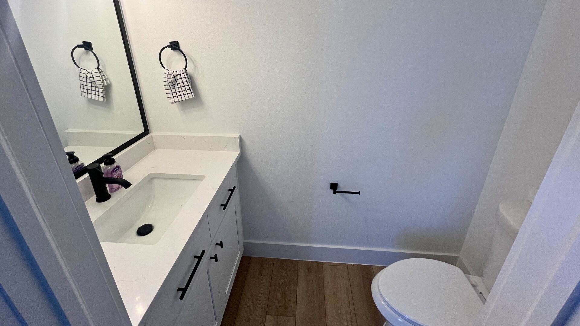 A small bathroom with a white sink, a mirror, a toilet, and a wooden floor.