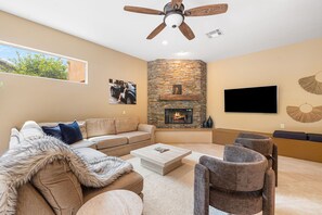 A modern living room with a sectional sofa, a fireplace, a ceiling fan, and a flat-screen TV. - Nightingale by Nest Villas | Lush La Quinta Oasis (La Quinta)