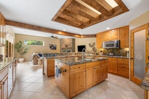 A spacious kitchen with a large island, wooden cabinets, a stone fireplace, and a modern microwave. - Nightingale by Nest Villas | Lush La Quinta Oasis (La Quinta)