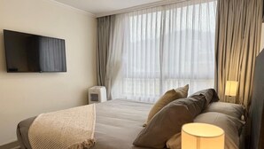 Quarto de hotel com cama, televisão na parede, abajur e janela com cortinas.