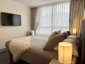 Ein Hotelzimmer mit Bett, Wandfernseher, Nachttischlampe und einem Fenster mit Vorhängen.