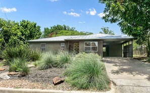 A single-story house with a metal roof, a covered garage, and a landscaped front yard with various shrubs and a rock. - The Nest | Charming 2BR Fredericksburg home with grill, full kitchen, and washer/dryer (Fredericksburg)
