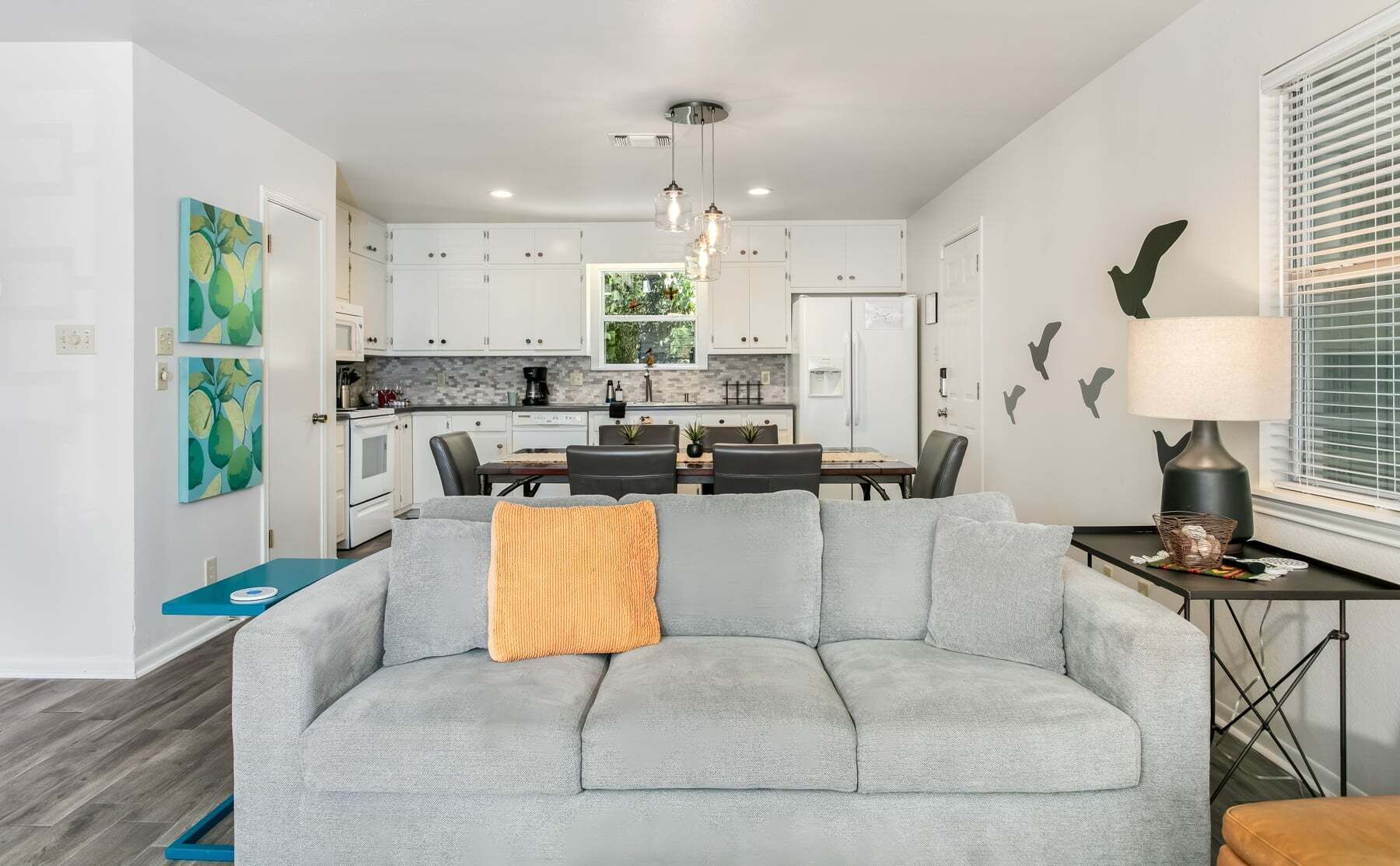 Ein modernes Wohnzimmer mit einem grauen Ecksofa, einem Essbereich mit Tisch und Stühlen, einer Küche mit weißen Schränken und einer Pendelleuchte.