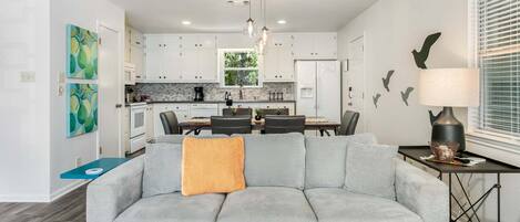 Una sala moderna con un sofá seccional gris, una zona de comedor con una mesa y sillas, una cocina con armarios blancos y una lámpara colgante.