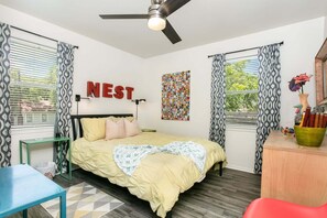 A bedroom with a bed, a desk, a chair, a ceiling fan, and a wall with a 'NEST' sign. - The Nest | Charming 2BR Fredericksburg home with grill, full kitchen, and washer/dryer (Fredericksburg)