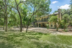 A two-story building with a balcony, surrounded by trees and a grassy yard. - Rio Ranchito | 3BR, 3BA river retreat with hot tub and fire pit (Stonewall)