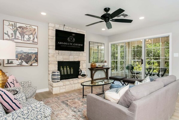 A living room with a fireplace, a ceiling fan, a large window, and a sofa. - Rio Ranchito | 3BR, 3BA river retreat with hot tub and fire pit (Stonewall)
