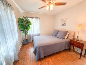 Un dormitorio con cama, mesita de noche, lámpara, ventilador de techo, ventana con cortinas y una planta en maceta.