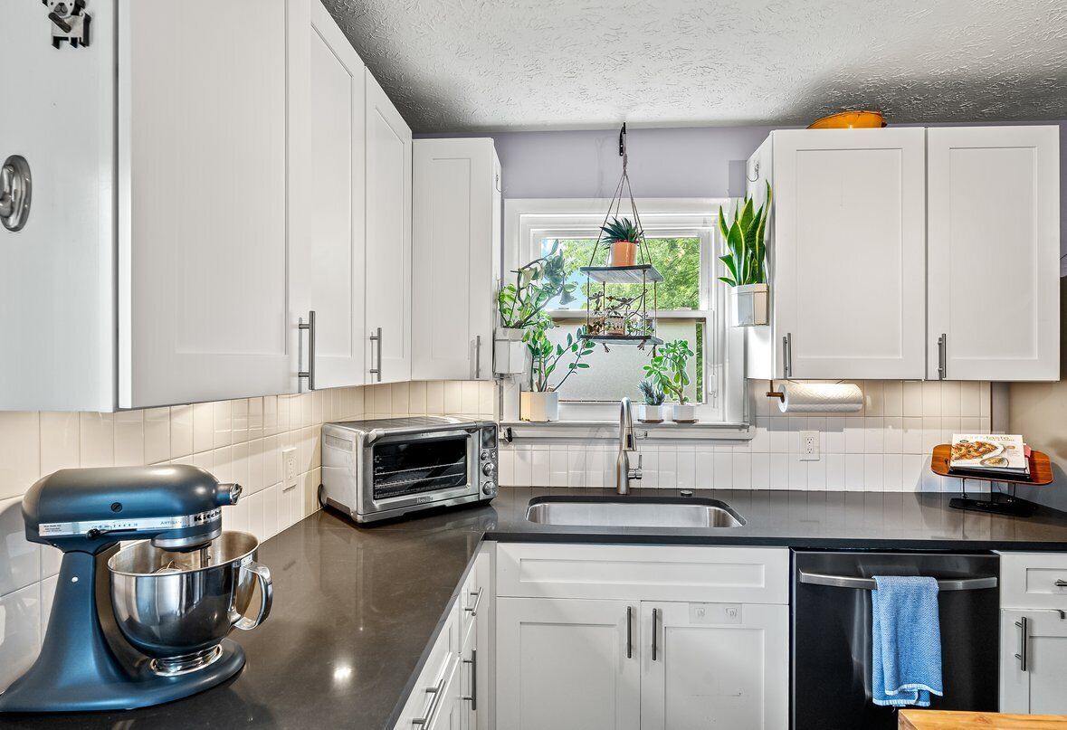 A modern kitchen with white cabinets, a black countertop, a stand mixer, a toaster, and a window with plants.