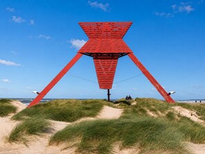 Eine rote Struktur mit einer dreieckigen Basis und flacher Spitze, gelegen an einem sandigen Strand mit Dünen und blauem Himmel.