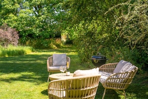 An outdoor seating area with wicker chairs and a table, surrounded by lush greenery. - Tudor style timber framed forest lodge - The Pink House (Bury St Edmunds)